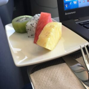 A small white plate with slices of kiwi, dragon fruit, watermelon, and pineapple is placed next to a laptop. A fork and knife are on a napkin beside the plate.