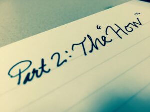 The image shows a close-up of a piece of lined paper with the handwritten text "Part 2: The 'How'". The handwriting is in cursive and written with a dark ink pen. The background is slightly blurred, focusing on the text.