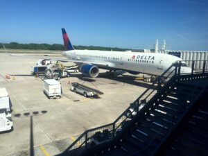 The image shows a Delta Airlines airplane parked at an airport gate. Ground crew members are servicing the aircraft, with various equipment and vehicles around it, including baggage carts and a fuel truck. The airplane is connected to the jet bridge, and there is a staircase in the foreground leading up to the terminal. The sky is clear and blue, and there is a runway and some greenery in the background.