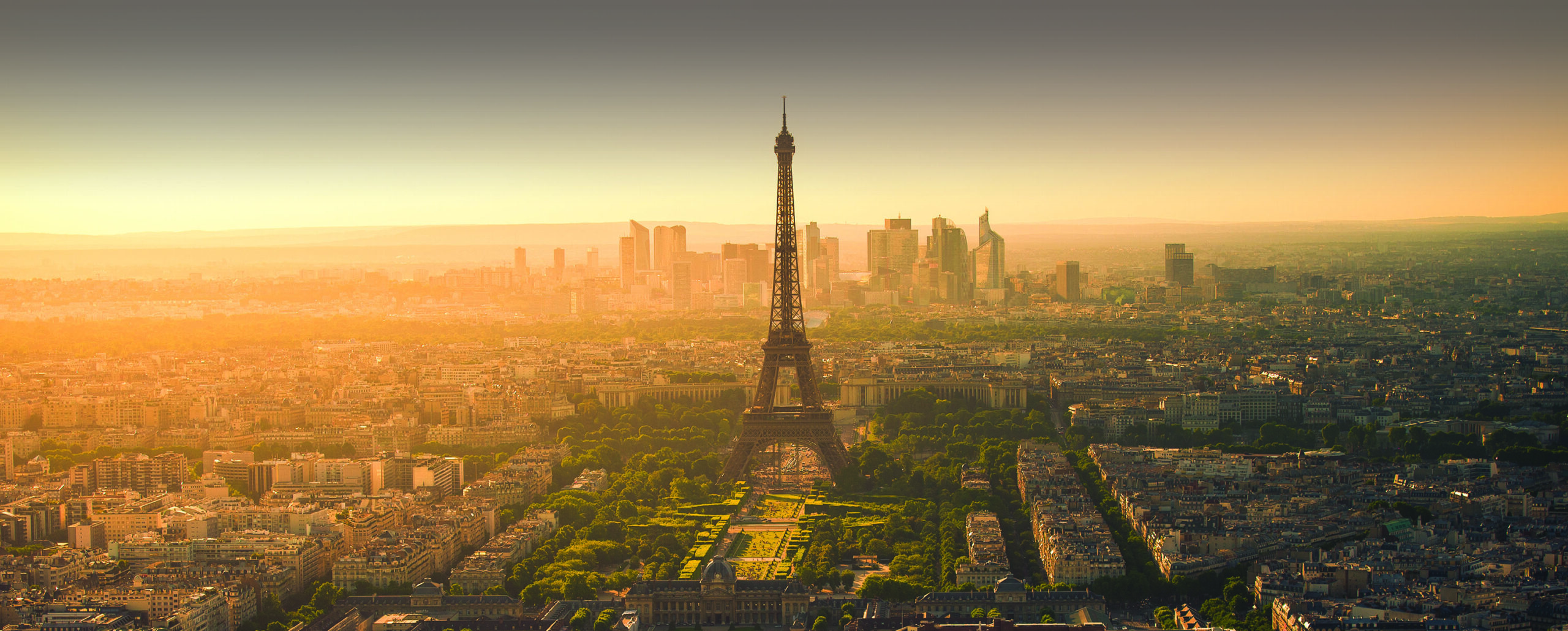 Alt text: A panoramic view of Paris at sunset, featuring the Eiffel Tower prominently in the center. The cityscape is bathed in a warm, golden light, with modern skyscrapers in the background and historic buildings and green spaces in the foreground.