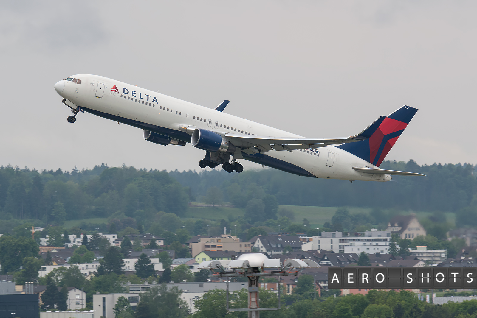 A Delta Air Lines aircraft is taking off, with its landing gear still extended. The plane is ascending against a backdrop of a cloudy sky and a landscape featuring trees and buildings. The image has the text "AERO SHOTS" in the bottom right corner.