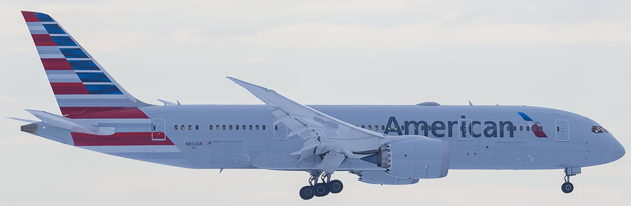 An American Airlines aircraft in flight, viewed from the side. The plane has a white fuselage with the American Airlines logo and a tail featuring red, white, and blue stripes. The landing gear is extended, indicating that the plane is either approaching for landing or has just taken off. The sky in the background is mostly clear with some light clouds.