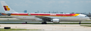 The image shows an Iberia Airlines aircraft on the tarmac at an airport. The airplane is white with a red and yellow stripe running along the fuselage and the Iberia logo on the tail. The background includes a clear sky and some airport infrastructure.